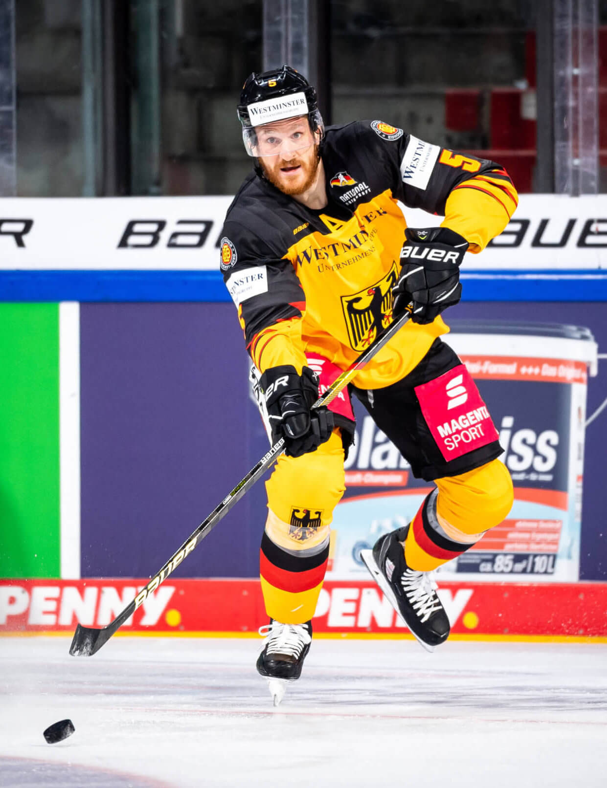 Das Bild zeigt den Eishockeyspieler Korbinian Holzer mit gelb und schwarzer Hose Trikot auf dem Eis. Er trägt einen schwarzen Helm mit Visier und hält einen Eishockeyschläger. Vor ihm liegt der schwarze Puck. Im Hintergrund sind Werbebanner und die Spielfeldbegrenzungen.